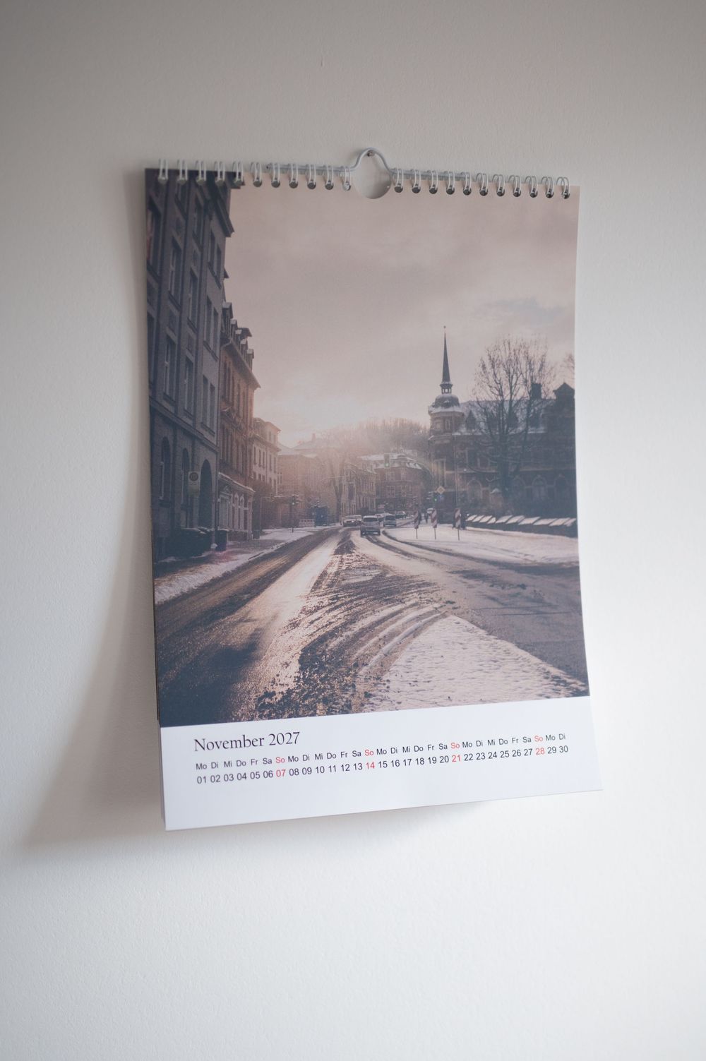 A wall calendar showing November 2027 with a snowy winter street scene featuring historic buildings and a church steeple.