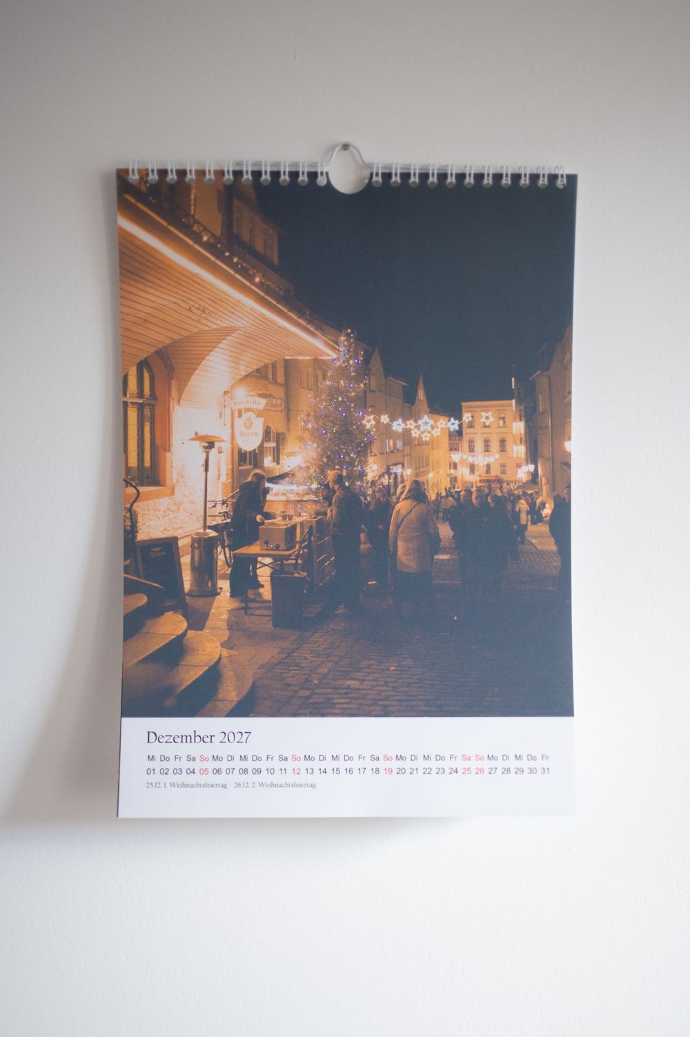 December 2027 wall calendar featuring a nighttime Christmas market scene with festive lights and crowds.