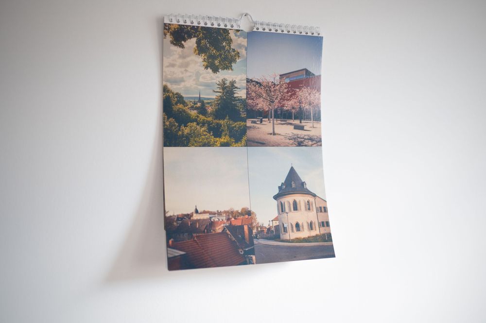 A spiral-bound wall calendar featuring four scenic outdoor photographs of buildings and landscapes hanging on a white wall.