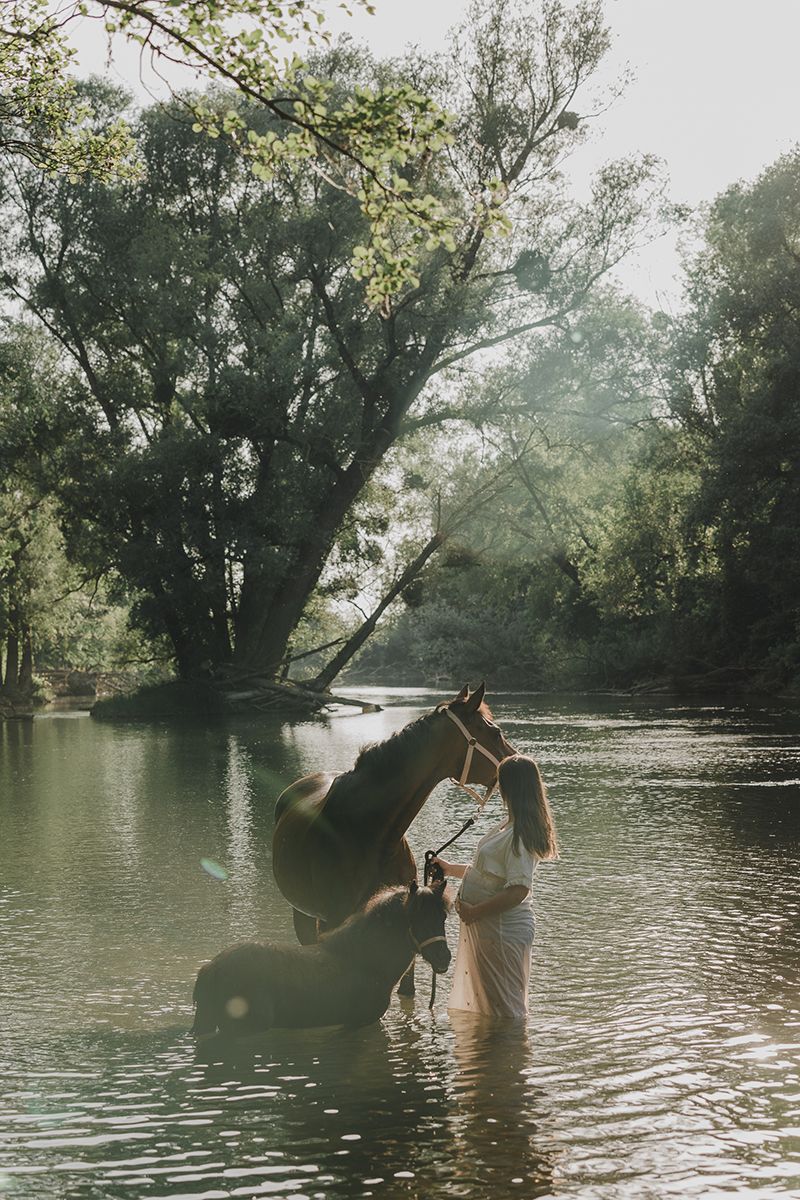 A woman in a white dress stands in a shallow river holding the reins of a dark horse, with another horse drinking beside her.