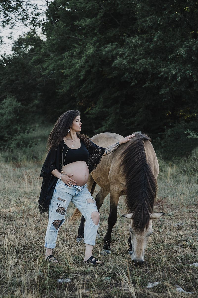 A pregnant woman with curly hair and ripped jeans gently touches a buckskin horse grazing in a grassy field at sunset.