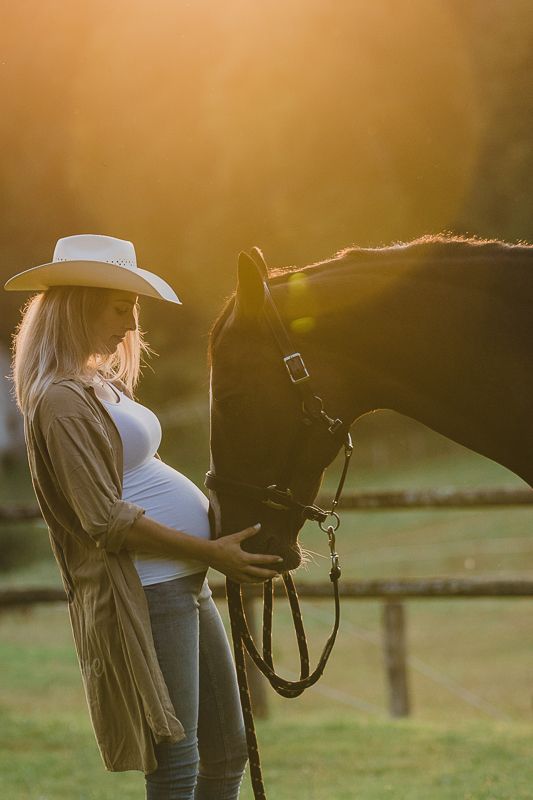 A pregnant woman in a white cowboy hat stands in golden sunset light, resting her hand on a dark horse's face.