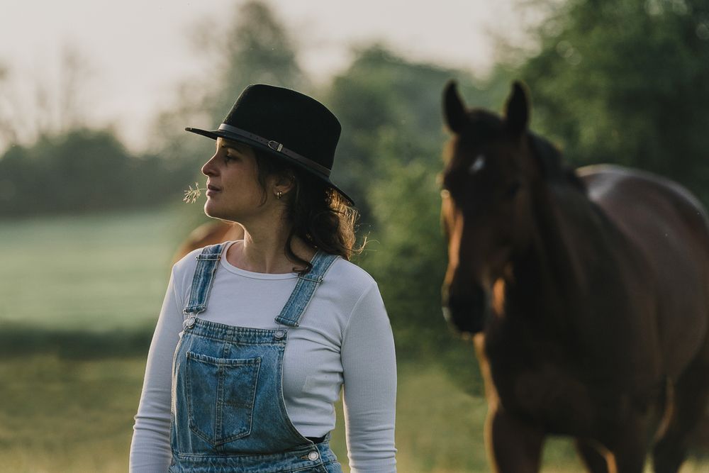 A woman in a black hat and denim overalls stands in a pasture with a brown horse behind her.
