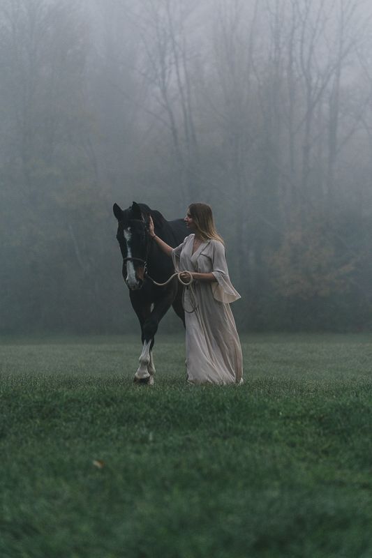 A woman in a flowing cream dress leads a black horse with a white blaze across a misty green field.