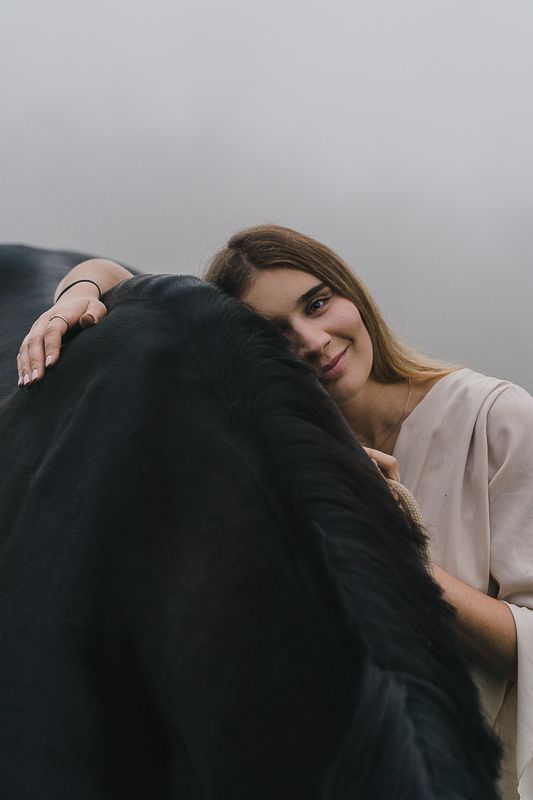 A smiling woman rests her head against a black horse's mane in a soft, hazy, dreamy portrait.