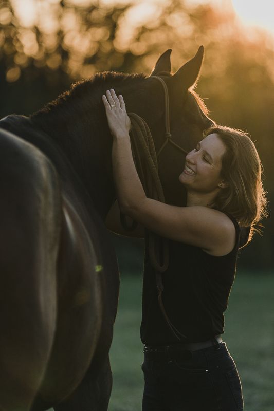 Smiling woman in a black top gently petting a dark brown horse in a golden sunset field.