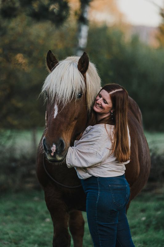 A smiling woman with long hair hugs a chestnut horse with a pale blonde mane in a green pasture.