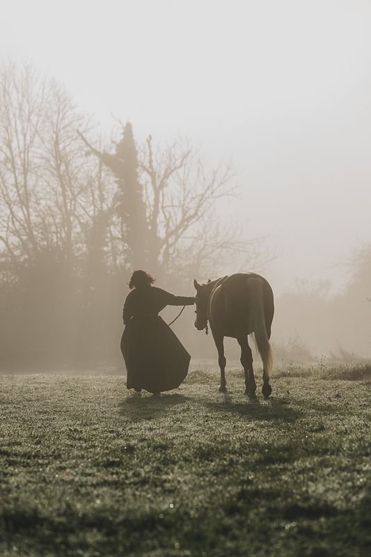 Woman in a long dark dress leading a horse across a misty, frosty meadow at sunrise.