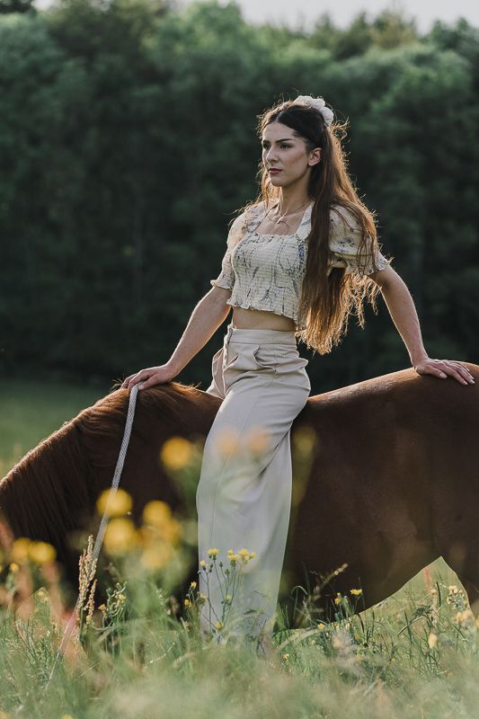 Young woman in a cream crop top and linen pants standing beside a chestnut horse in a wildflower meadow.