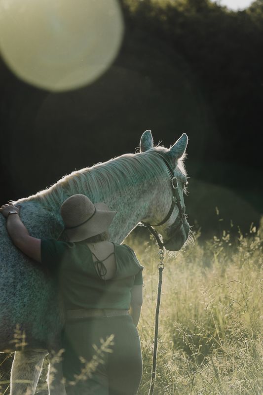 Woman in a wide-brimmed hat hugging a dappled gray horse in tall grass with warm backlit sun flare.
