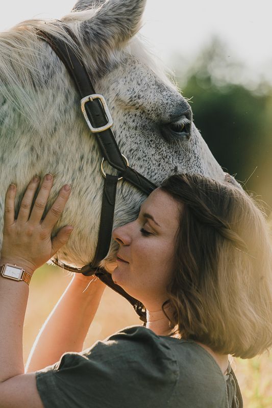 Woman in a green t-shirt resting her face lovingly against a gray horse in warm golden light.