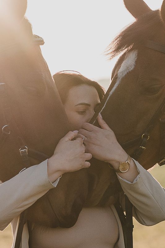 Woman in a beige jacket tenderly cradling the faces of two horses in soft, warm sunlight.