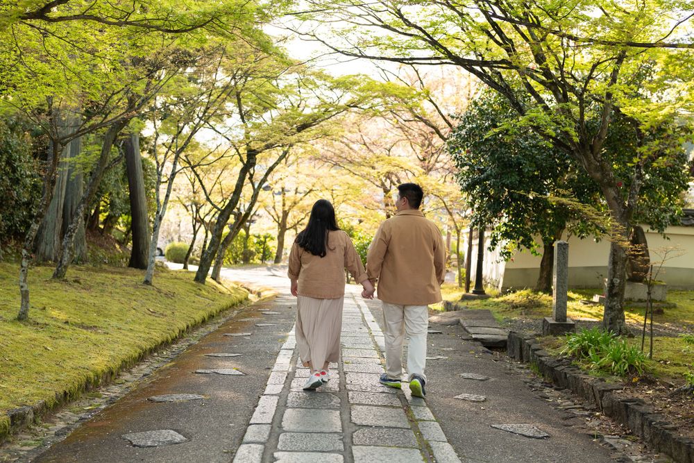 Couple in traditional Japanese kimono and haori holding hands in front of a historic temple with cherry blossoms and stone lanterns.