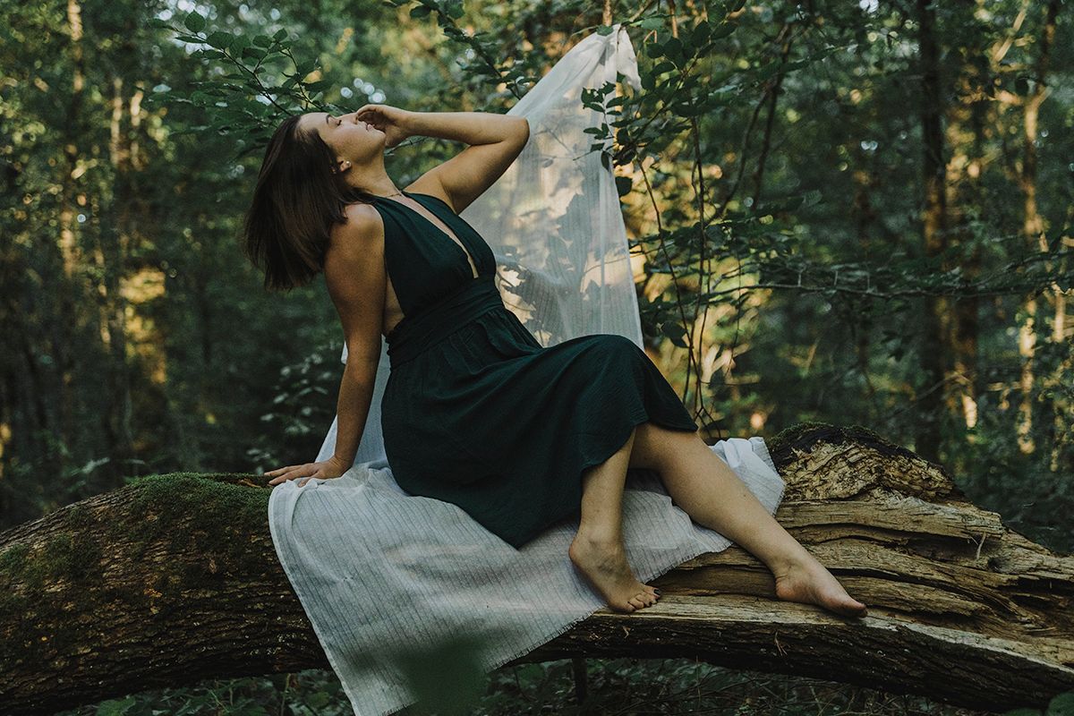 Woman in a dark green dress sits on a mossy fallen log in a sunlit forest with a white fabric draped behind her.
