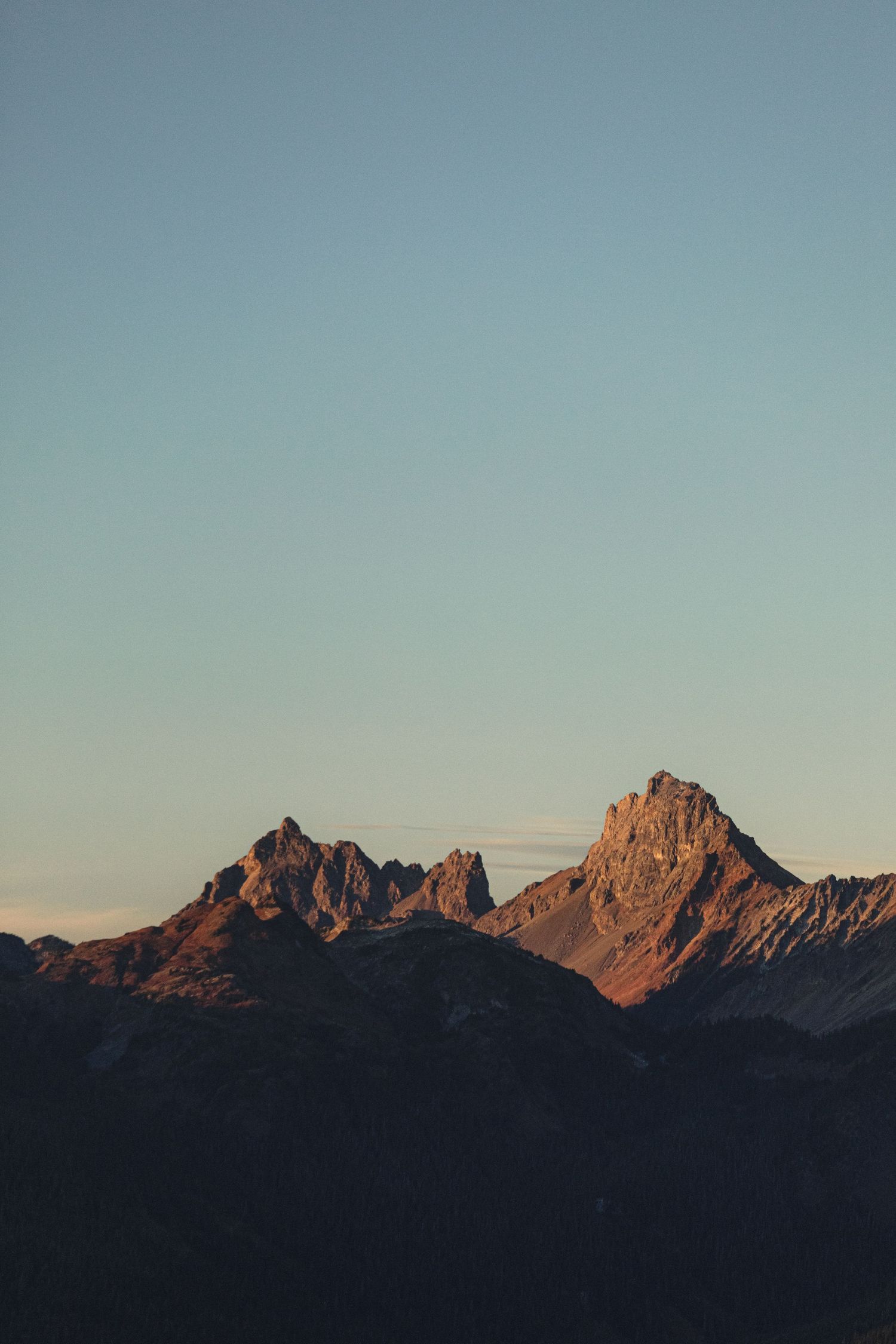 Rugged mountain peaks glowing in warm orange sunset light against a clear pastel blue sky, with dark shadowed ridges in the foreground.