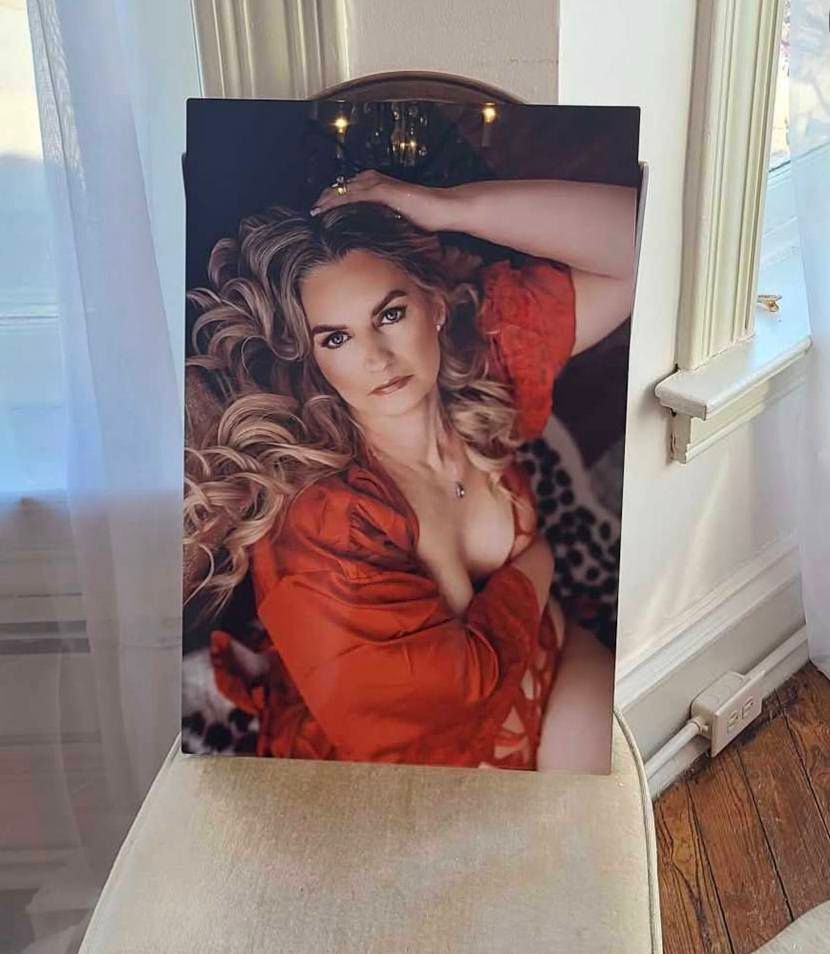 Framed boudoir portrait of a woman with long wavy blonde hair in a red lace robe, displayed on a cushioned chair beside a sunlit window.