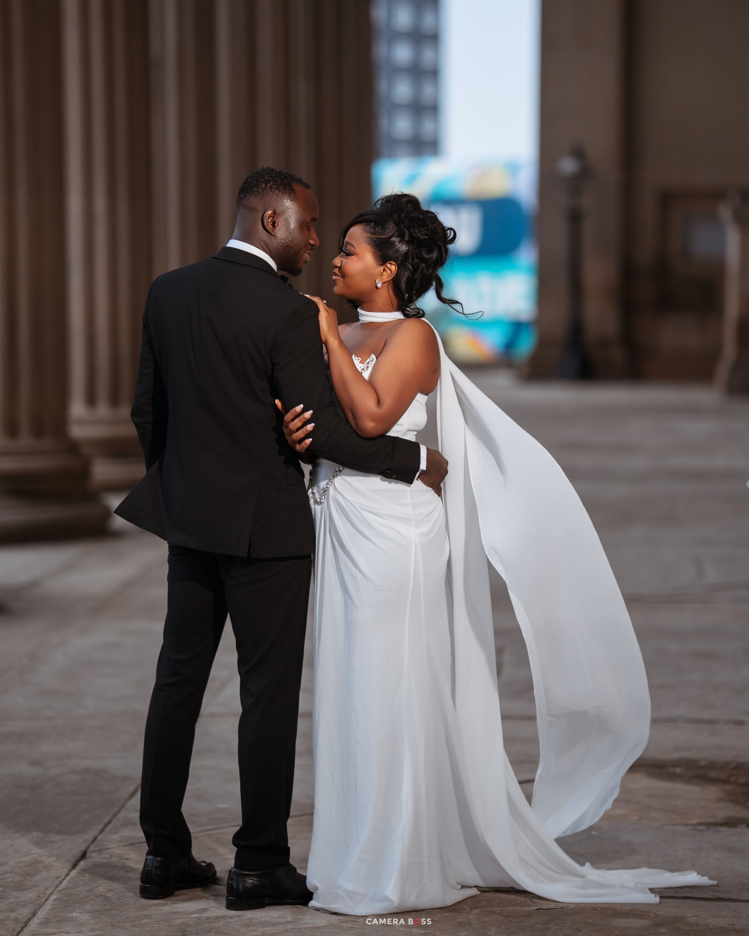 A couple in wedding attire share a romantic moment, the bride in a white gown with flowing cape and groom in a black suit.