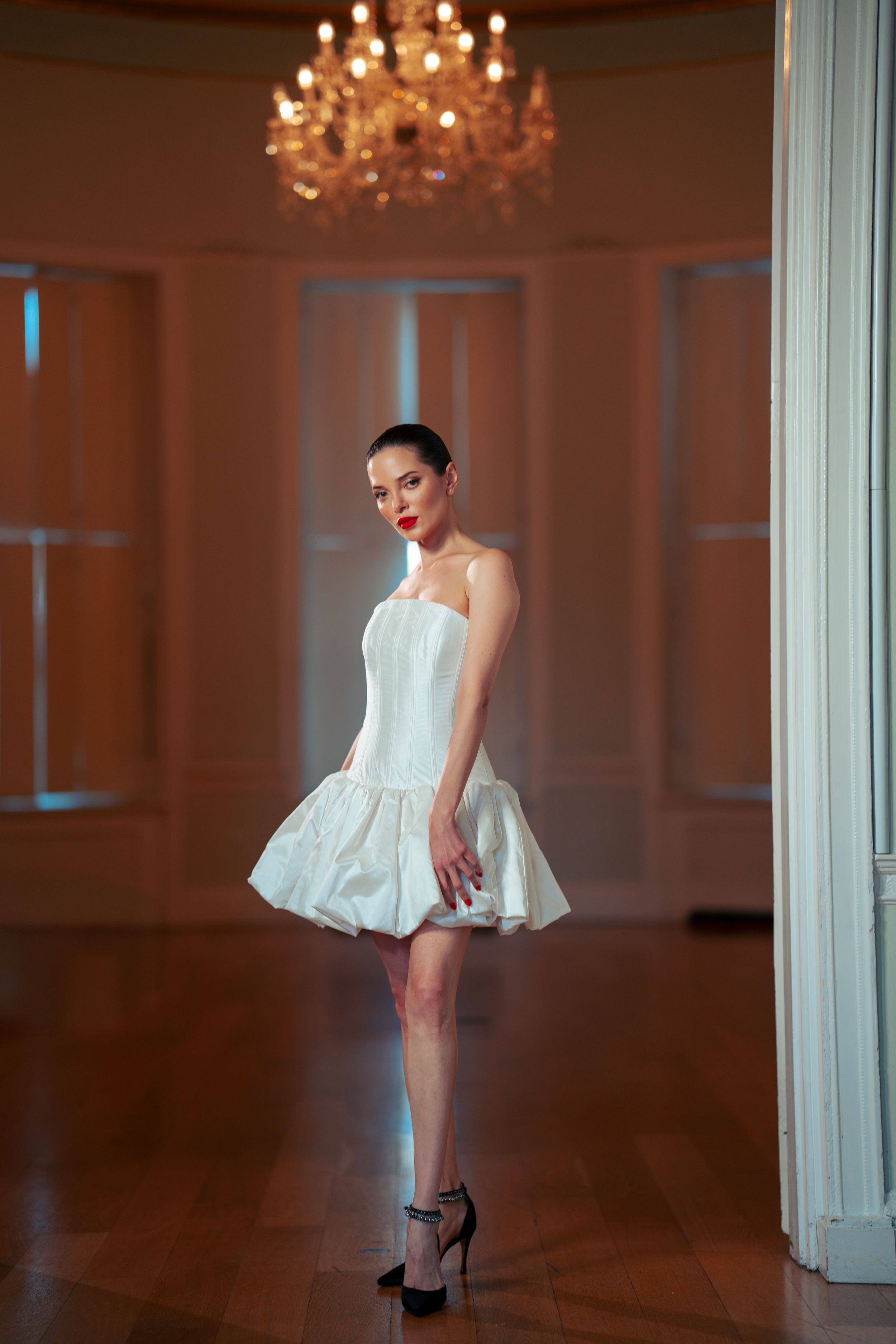 A lady in a short white bubble dress and high heels poses elegantly in an elegant room with a crystal chandelier.