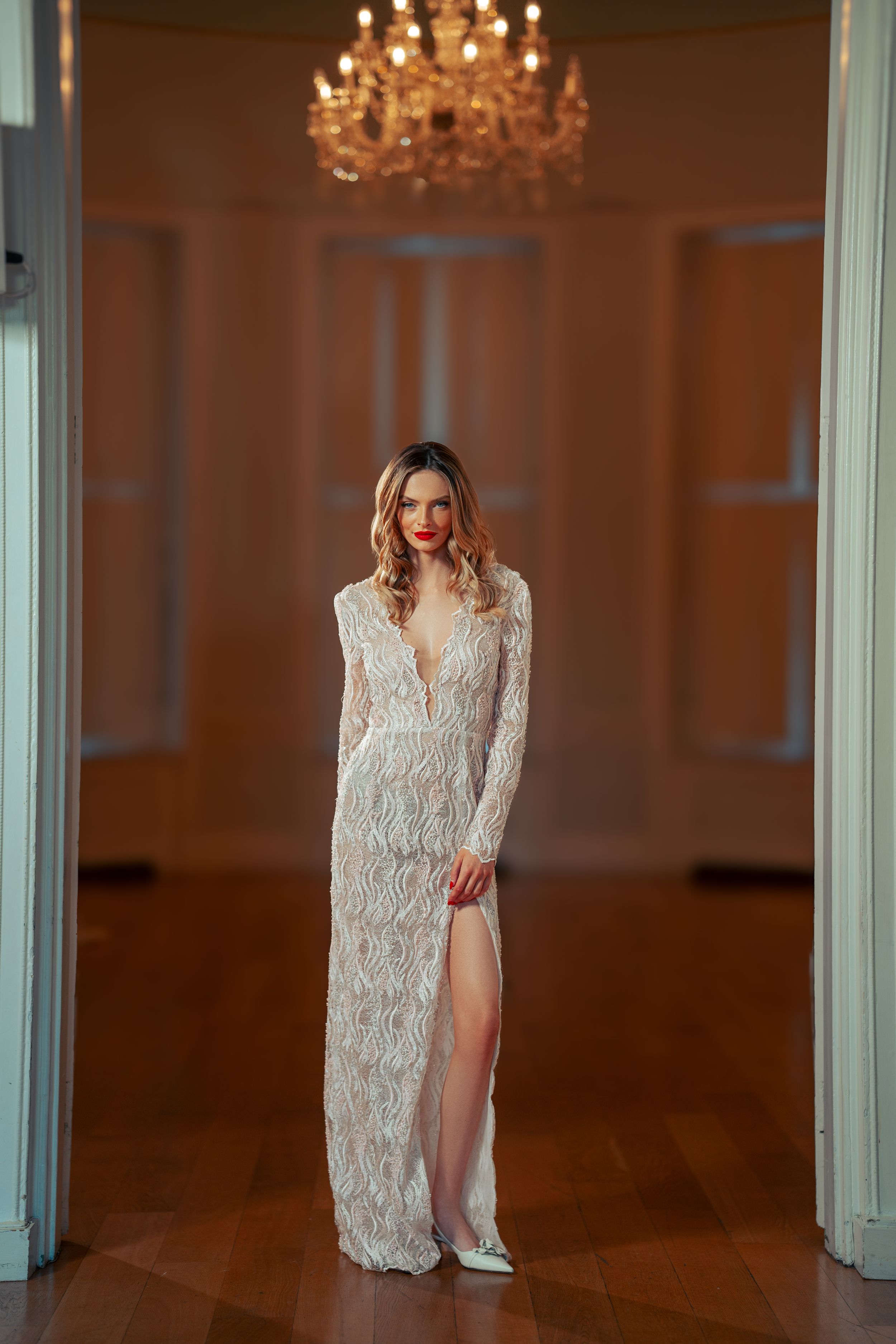 Elegant white lace wedding dress with plunging neckline and thigh-high slit under crystal chandelier in formal room.