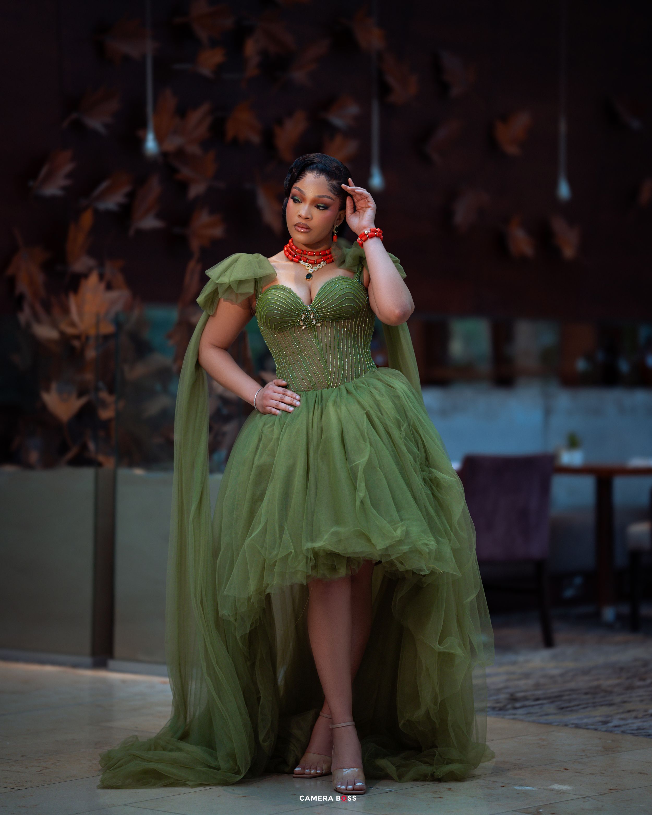 A model in a flowing olive green high-low gown with ruffled tulle skirt poses elegantly in an outdoor setting.