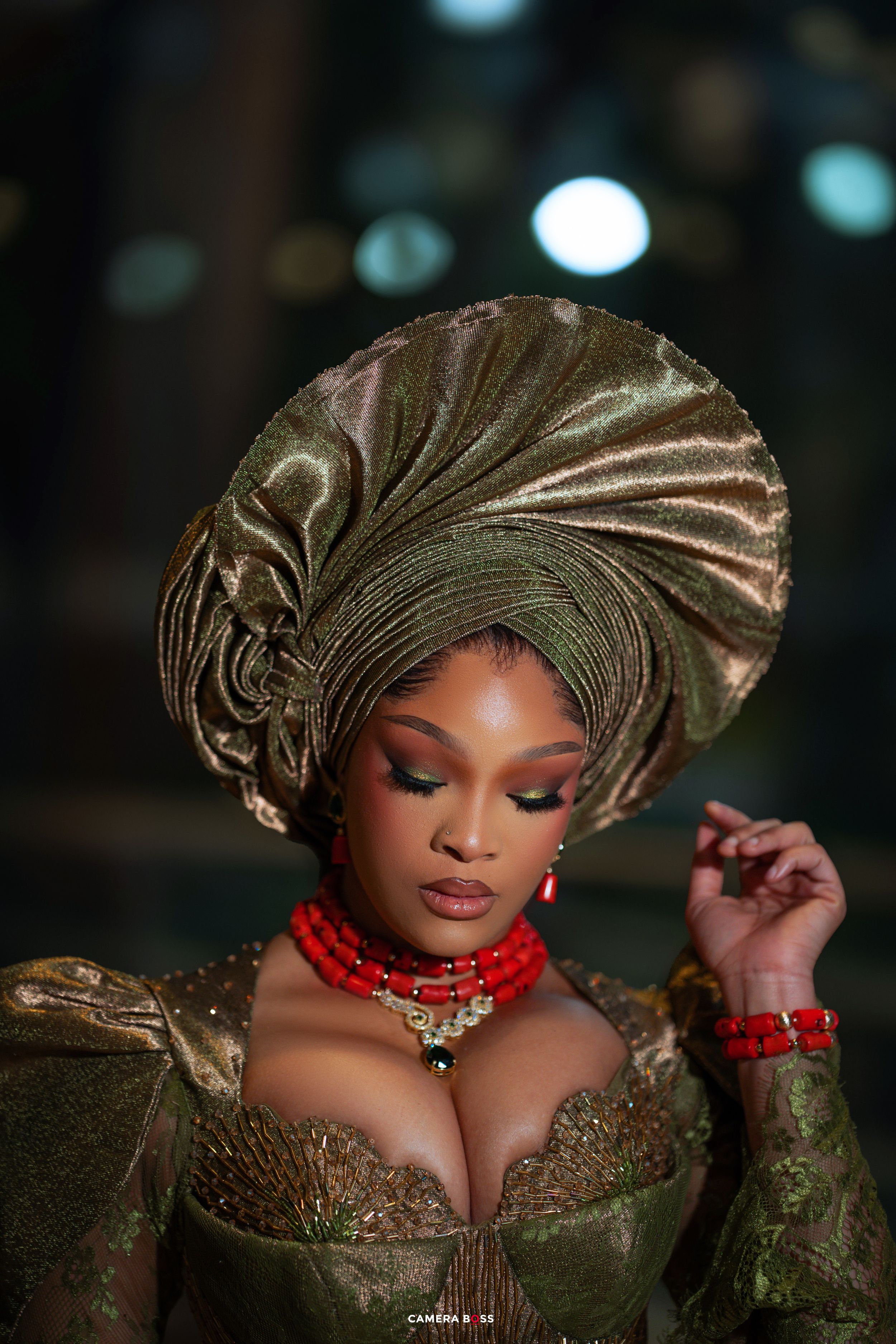A woman in traditional Nigerian attire with gold gele headpiece and red necklace poses elegantly against dark background.