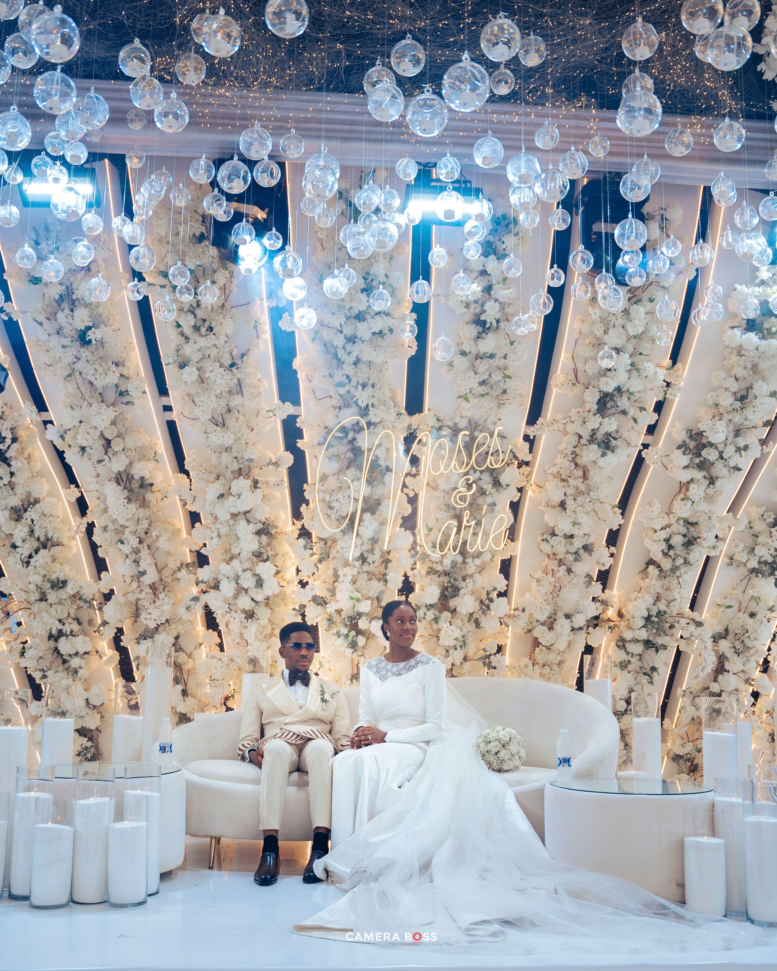 An elegant wedding ceremony setup with hanging glass orbs, white pillars and floral decorations against a light backdrop.