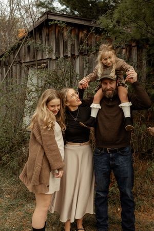 A happy family of four poses outdoors near a rustic barn, with a young girl sitting on her fathers shoulders.