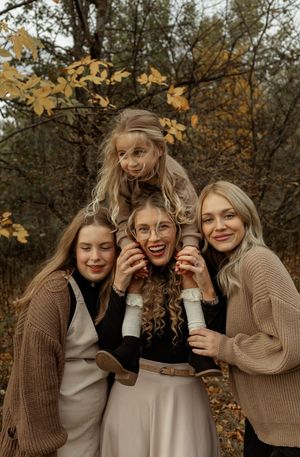 Four blonde women pose together outdoors in autumn, smiling warmly among golden fall foliage.