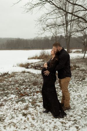 Couple embracing in snowy winter landscape, pregnant woman in black dress, man in dark jacket, bare trees and frozen lake behind them.