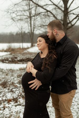 Couple in black outfits embracing outdoors in snow, woman visibly pregnant, man kissing her head, winter maternity photo session.