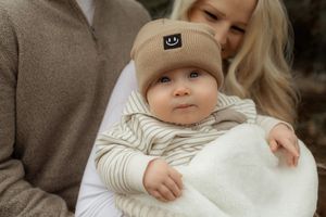 Adorable blue-eyed baby in beige smiley face beanie wrapped in white blanket held by parents.