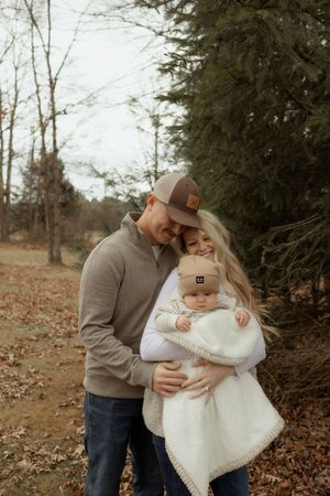 A couple tenderly embraces outdoors in autumn, holding their baby wrapped in a white blanket near evergreen trees.