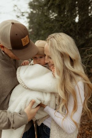 A couple smiles while holding their baby wrapped in a white blanket outdoors among trees.