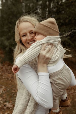 Smiling blonde mother holding her baby dressed in beige outfit and knit hat outdoors in autumn forest.