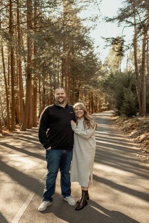 Smiling couple posing on a tree-lined road in a forest, man in black jacket and jeans, woman in cream sweater dress.