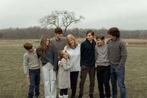 Large family group portrait outdoors in a field on a cloudy day, wearing casual neutral tones, smiling together.