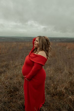 Pregnant woman in red off-shoulder dress standing in a field, cradling her baby bump on an overcast day.