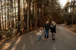 Family of three walking hand-in-hand down a sunlit forest road surrounded by tall pine trees in autumn.