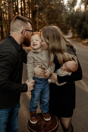 Family portrait with parents kissing toddler boy outdoors on a forest road during golden hour.