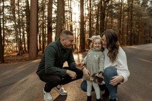 Family of three with a toddler and small dog posing on a forest road surrounded by tall trees in warm autumn light.