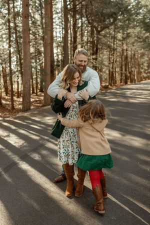 A man hugs a woman and young girl on a tree-lined road in a warm family embrace outdoors.