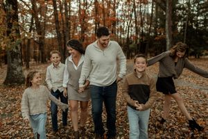 Family of six walking through autumn woods, laughing and holding hands among fallen leaves.