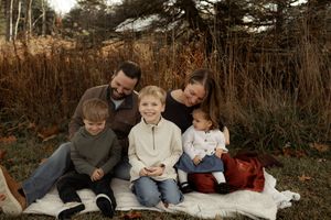 Family of five sitting on a blanket outdoors in autumn, smiling together with tall golden grasses in the background.