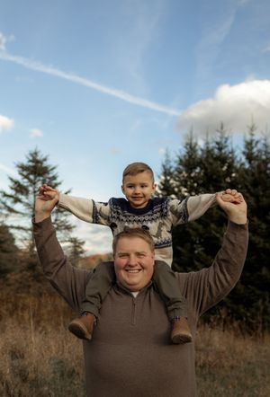 A smiling man holds a happy young boy on his shoulders outdoors near evergreen trees under a blue sky.