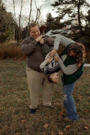 Two parents playfully flip their laughing child upside down outdoors in a yard with autumn leaves.