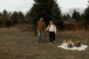 Family holding hands in autumn field with evergreen trees, two young children playing on white blanket nearby.