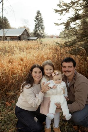 Family of four poses outdoors in autumn field with rustic barn, parents kneeling with two young daughters.