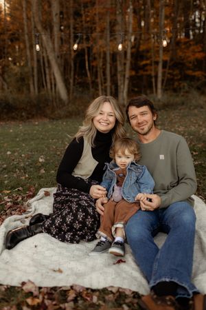 Family of three sitting on a blanket outdoors in autumn, smiling warmly with fall foliage and string lights behind them.