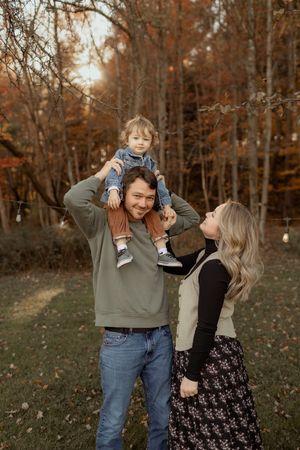 A happy family poses outdoors in fall, dad holds toddler on shoulders while mom smiles, surrounded by autumn foliage.
