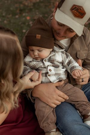 Family with baby in plaid shirt and brown beanie hat being held by parents in autumn outdoor setting.